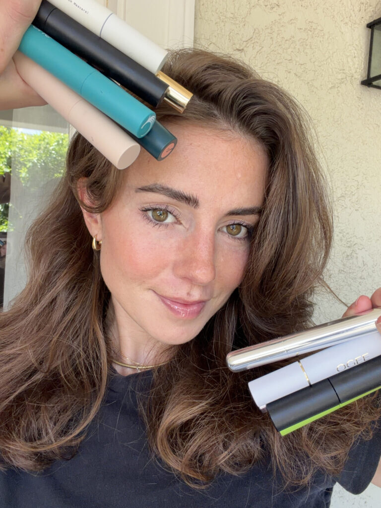 12 Best Natural And Organic Mascara Brands (2026 Review) A woman with wavy brown hair holds several mascara tubes near her face and head, smiling slightly at the camera.