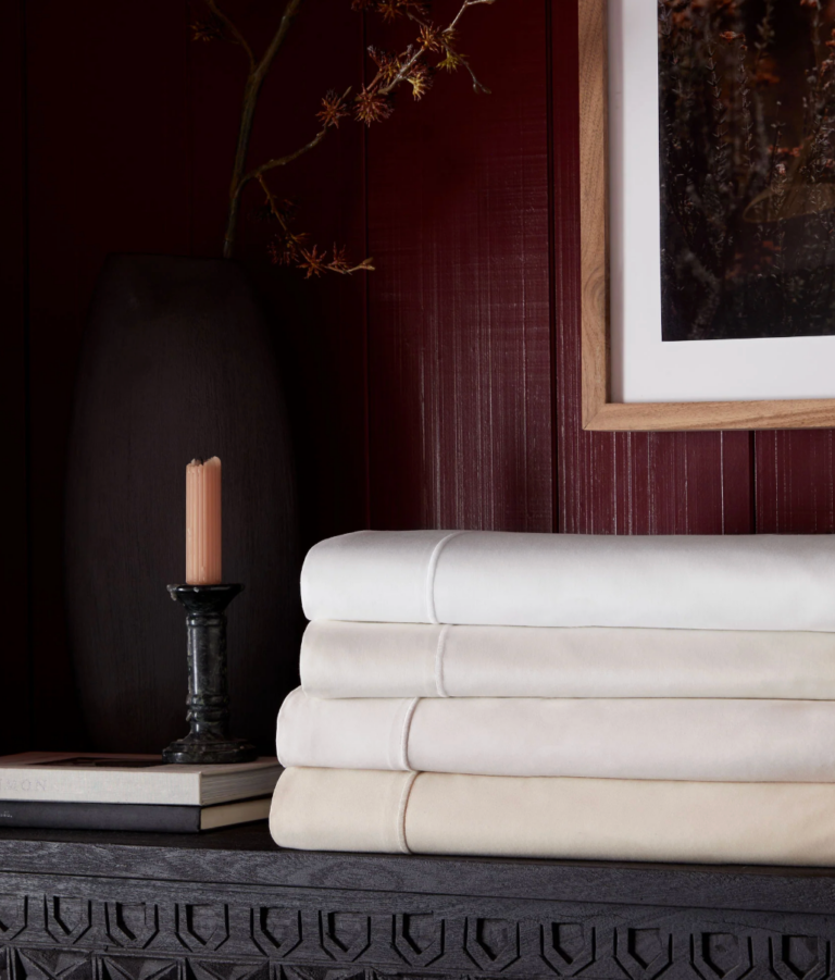 Three neatly folded sheets in light neutral colors sit on a dark wooden surface beside a candle, books, a vase with branches, and a framed picture on a red wall.