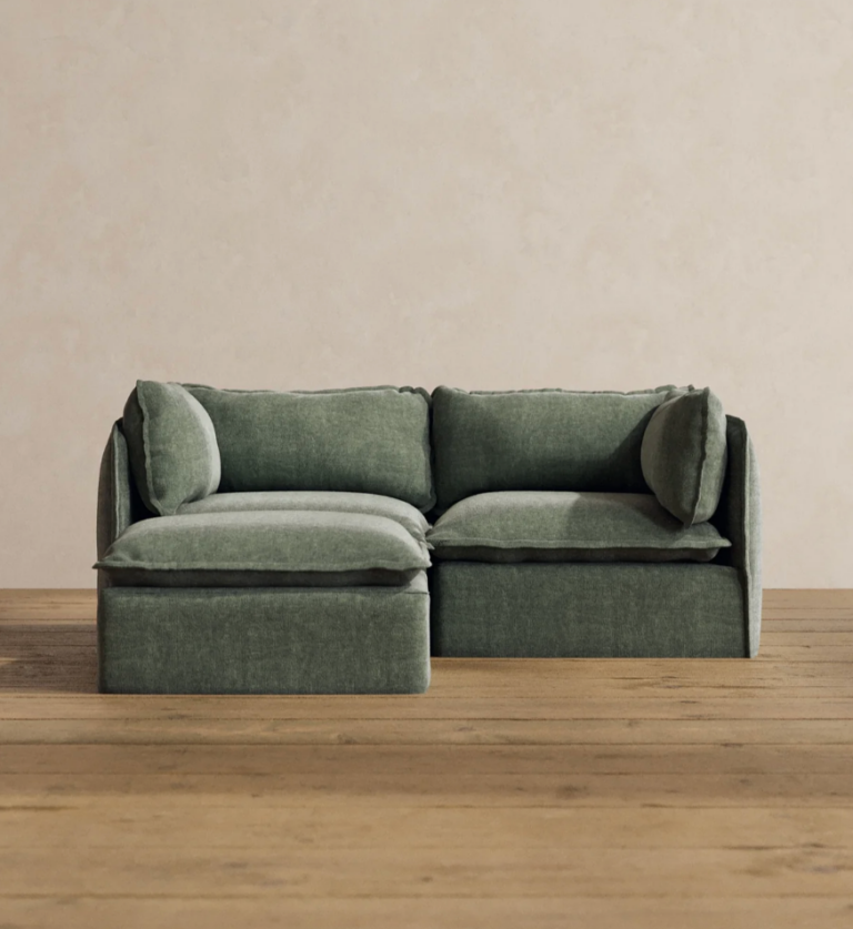 A green sectional sofa with two seat cushions, two back cushions, and two side pillows on a wooden floor against a beige wall.