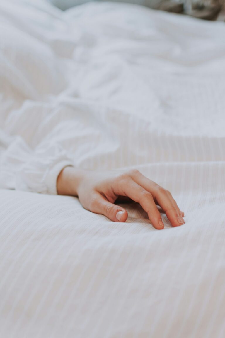A person's hand resting on a white bed sheet, partially covered by a white sleeve.