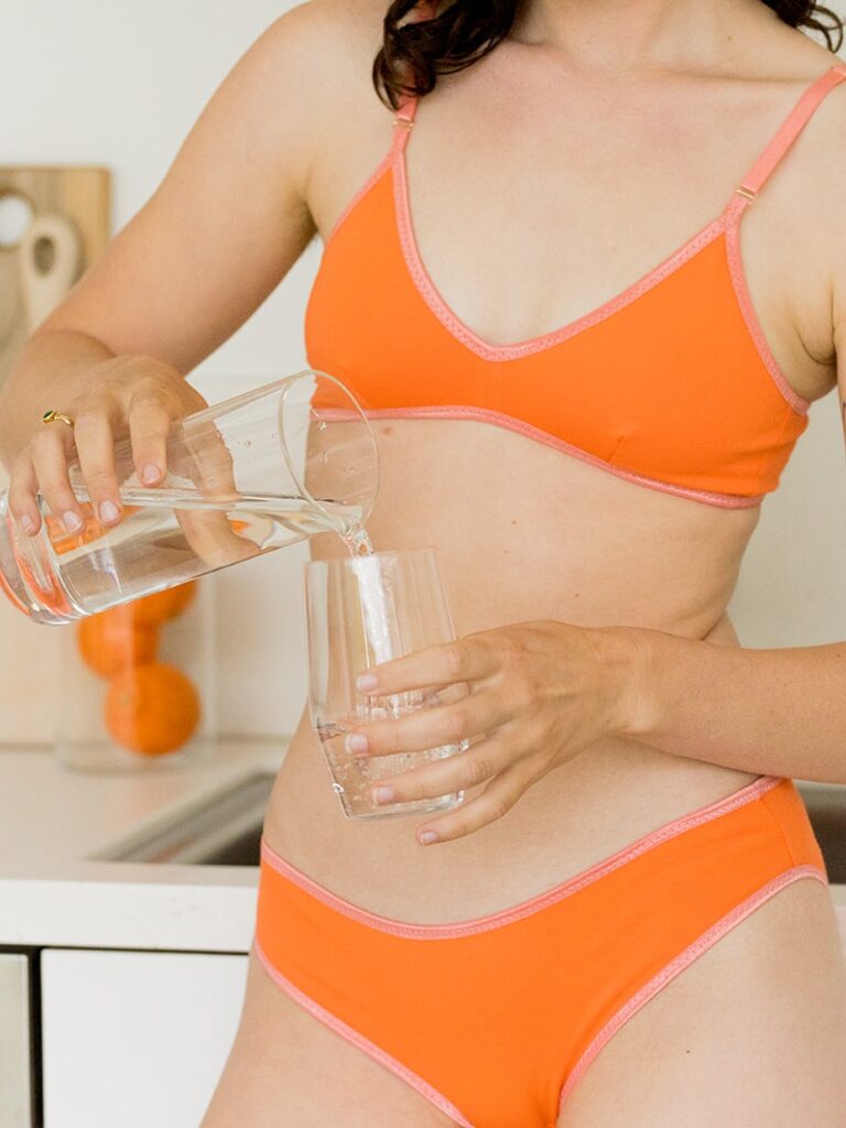 A woman in an orange bra and underwear pours water from a glass pitcher into a glass in a kitchen.