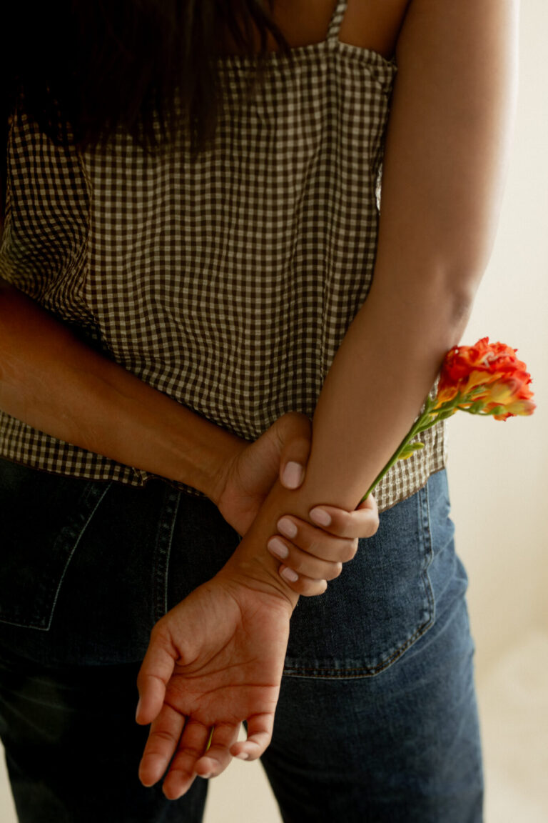 A person in a checkered top and jeans holds an orange carnation behind their back with both hands.