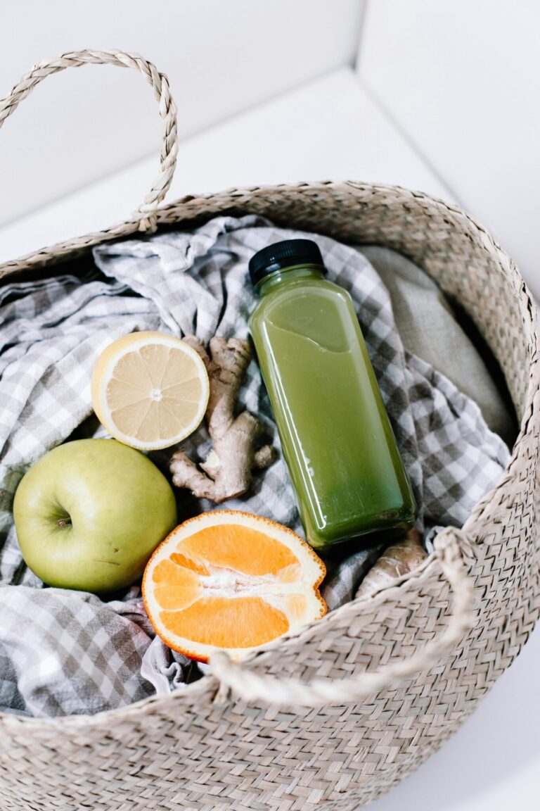A woven basket contains a green juice bottle, a green apple, half a lemon, half an orange, ginger root, and a gray gingham cloth.