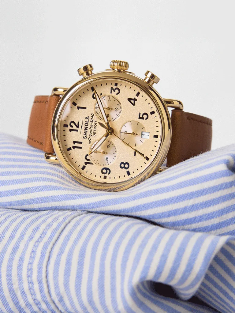 10 Best American-Made Watch Brands For 2026 A gold chronograph wristwatch with a brown leather strap rests on a folded blue and white striped shirt.