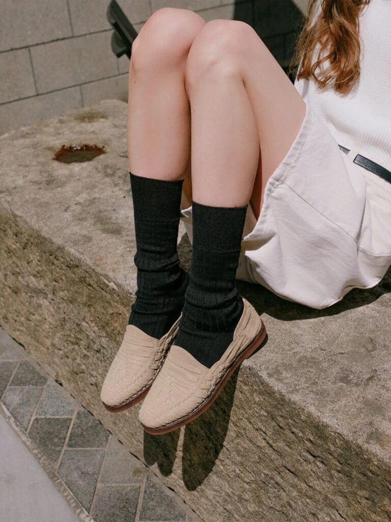 5 huaraches éticos y sandalias inspiradas en huarache (2026) A person sits on a stone ledge, wearing a white skirt, long black socks, and beige huarache sandals. Their hands rest gently on their lap.