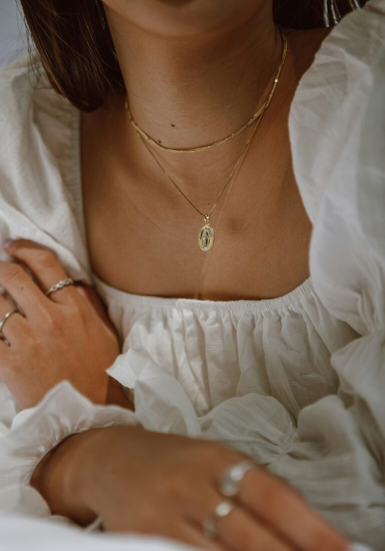 11 Best Places To Buy Affordable Vintage Jewelry Online A person wearing a white textured blouse, layered necklaces with a pendant, and silver rings on fingers.