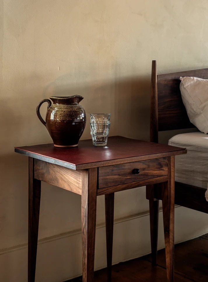 15 Best American Made Furniture Brands In 2026 A brown ceramic pitcher and a clear glass sit on a wooden bedside table next to a bed with white bedding against a beige wall.