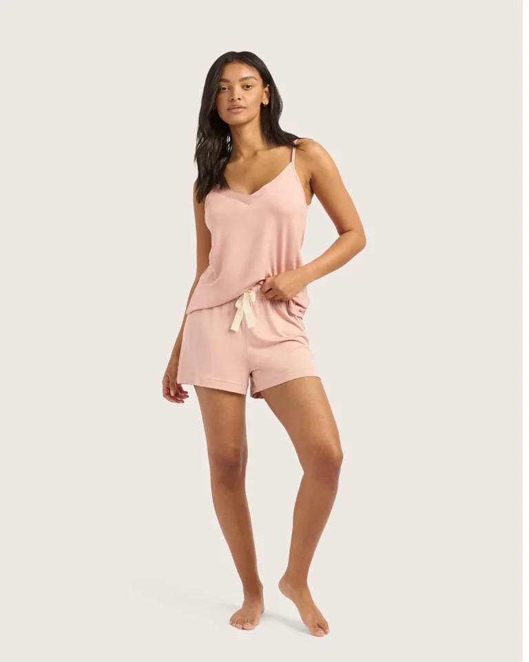 7 Bamboo Pajamas For Women (2026) A woman stands barefoot wearing a light pink camisole and matching shorts with a drawstring, posed against a plain background.