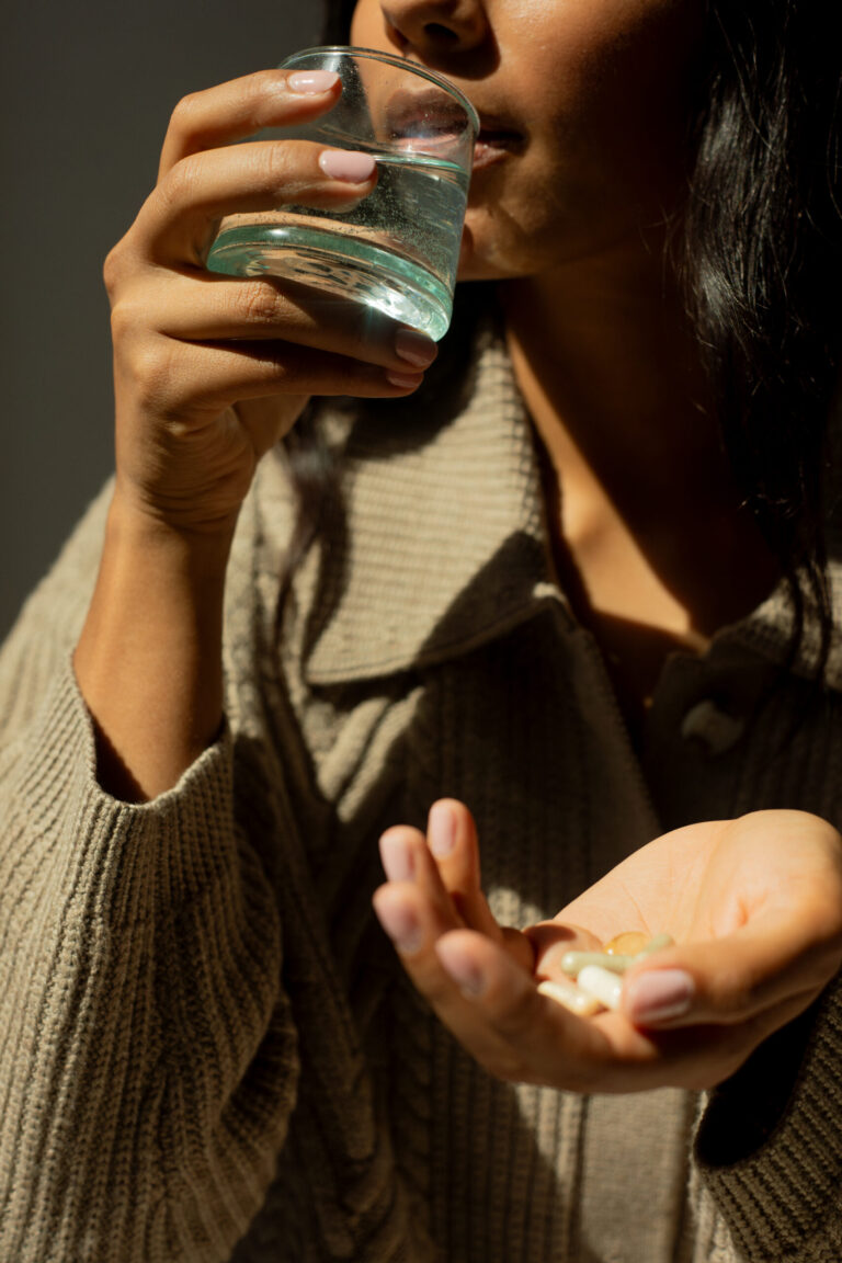 8 Best Hair Growth Supplements And Vitamins (2026) A person holds several capsules in one hand and lifts a glass of water to their mouth with the other hand, wearing a beige sweater.