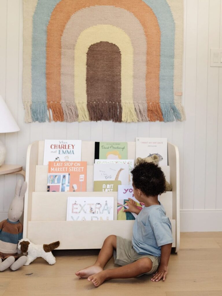 9 Best Montessori Bookshelves For Early Readers A child sits on the floor, reaching for a book from a wooden bookshelf filled with children