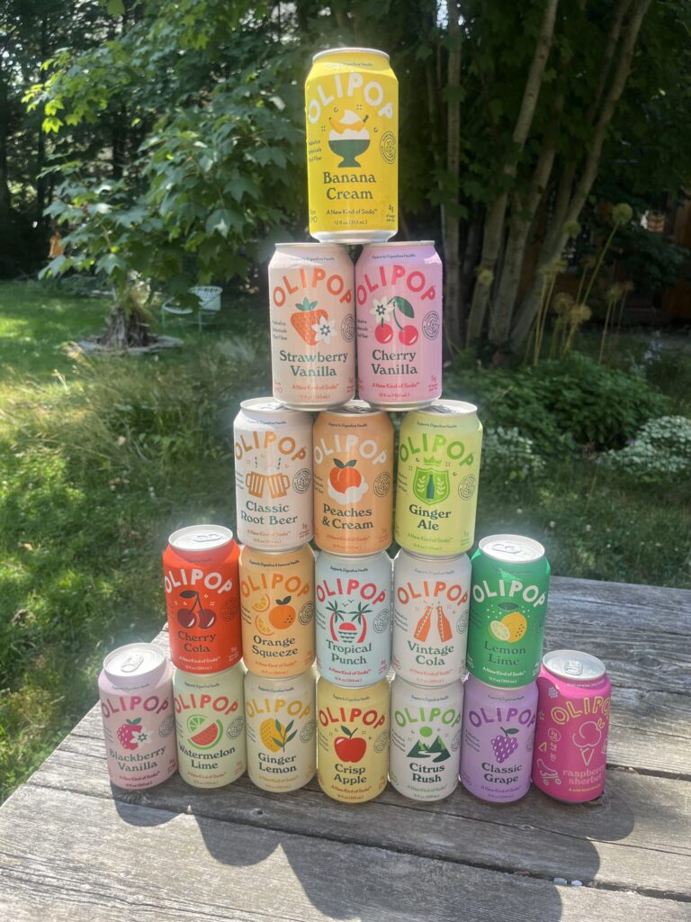 I Tried Every Flavor Of Olipop And Ranked Them (2026) A pyramid of sixteen colorful OLIPOP soda cans in various flavors is displayed on a wooden table outdoors, with trees and greenery in the background.