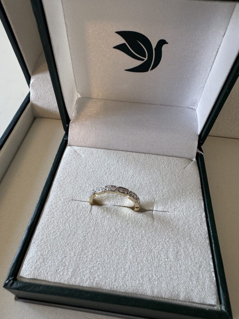 Por qué elegí diamantes cultivados en laboratorio para mi alianza de boda A gold ring with several small diamonds is displayed in a white cushioned jewelry box with a green bird logo on the inside lid.