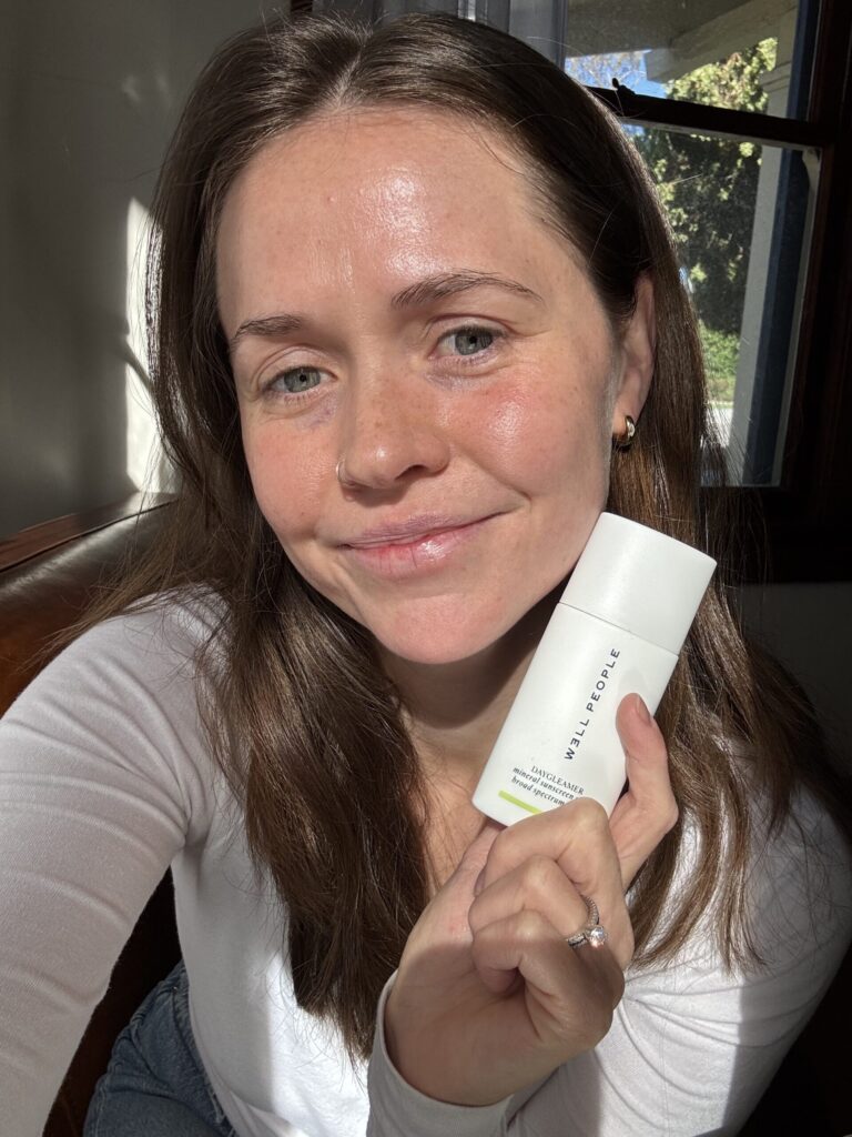 ¿Es el protector solar Daygleamer de Well People un producto imprescindible para el cuidado de la piel? Revisamos Person smiling and holding a white skincare product bottle indoors near a window.
