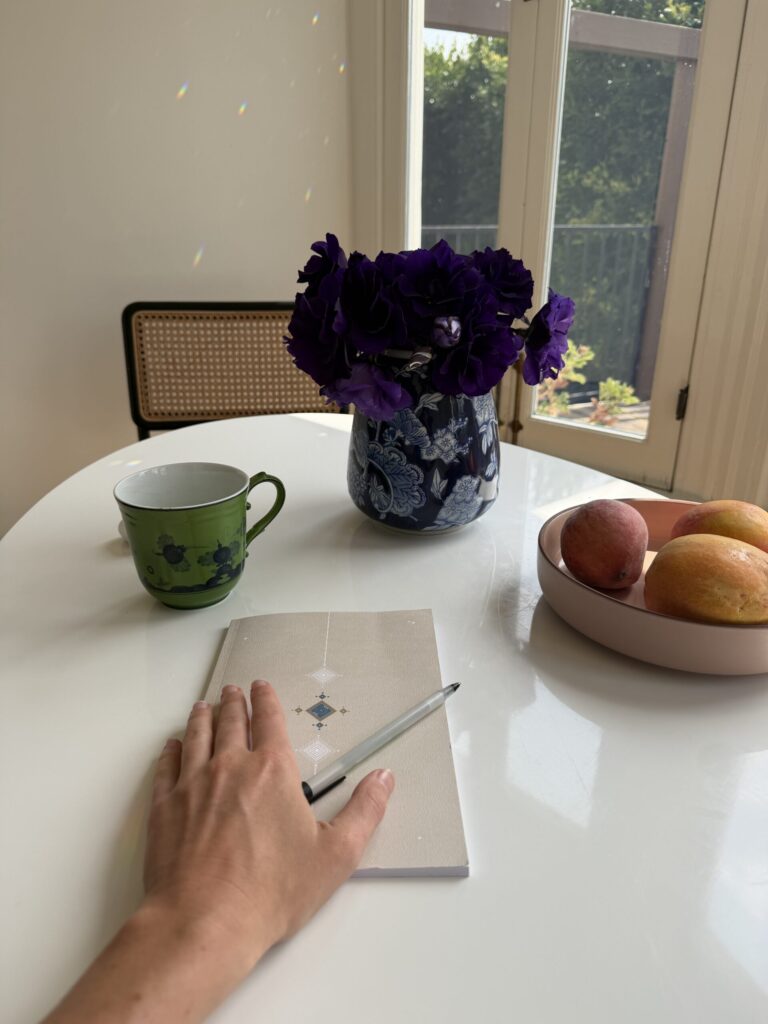 ¿Hacer «páginas matutinas» durante 30 días me hará más creativo? A hand rests on a closed notebook with a pen on a white table, next to a green mug, a vase of purple flowers, and a bowl of peaches and a mango.
