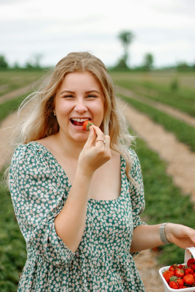 ¿Qué es la estética Cottagecore? Y por qué nos encanta A person with long blonde hair in a green floral dress smiles and holds a strawberry to their mouth while standing in a strawberry field. They have a container of strawberries in their other hand.
