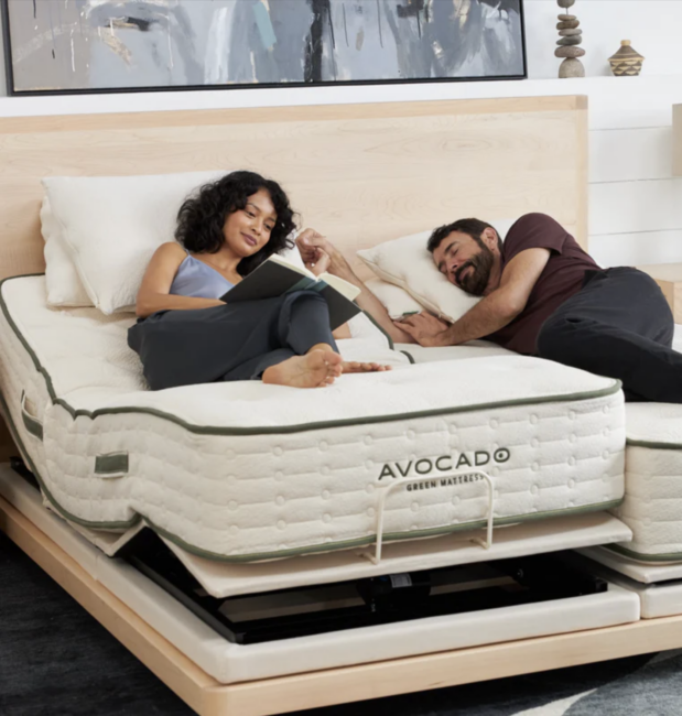9 Split King Mattresses Made With Nontoxic Materials A woman reads on an adjustable Avocado mattress while a man sleeps beside her on a separate section of the split bed in a modern bedroom.